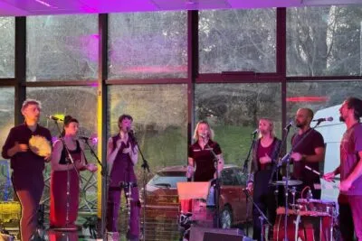 A live music performance in a bright, modern space with large windows showing bare tree branches outside. A group of seven vocalists, three men and four women, stand in a line holding microphones, performing energetically on stage under pink and purple stage lights.