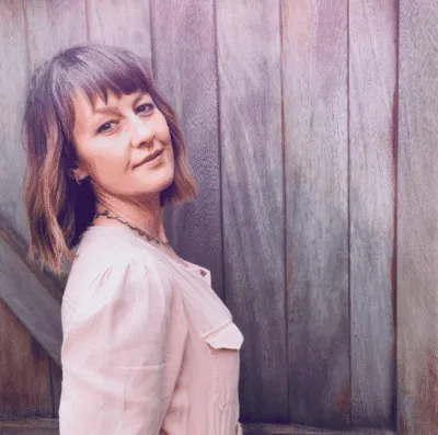 Emily Davies with light brown hair in light pink jacket standing against wooden background, looking thoughtfully to her right, soft natural light.