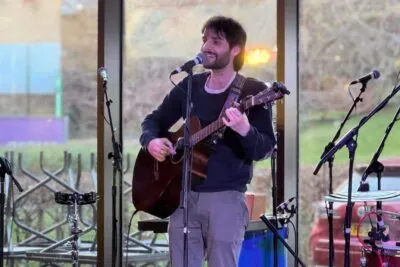 Jonah Corren singer-songwriter plays an acoustic guitar and sings into a microphone in a daytime indoor setting with large windows behind him. The natural light outside contrasts with the warm indoor lighting highlighting the performer.