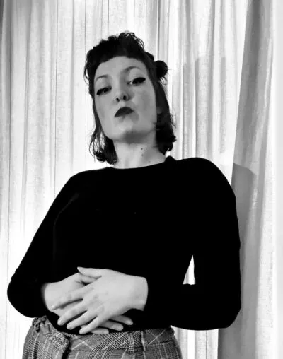 Black-and-white portrait of Moya Silk posing confidently against curtain backdrop, wearing dark top and patterned trousers, expressive facial expression.