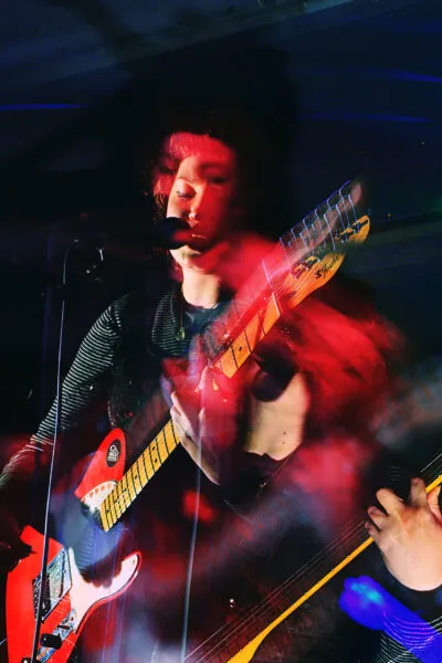 Close-up live image of Moya Silk passionately playing guitar and singing, surrounded by deep red and blue stage lighting hues.