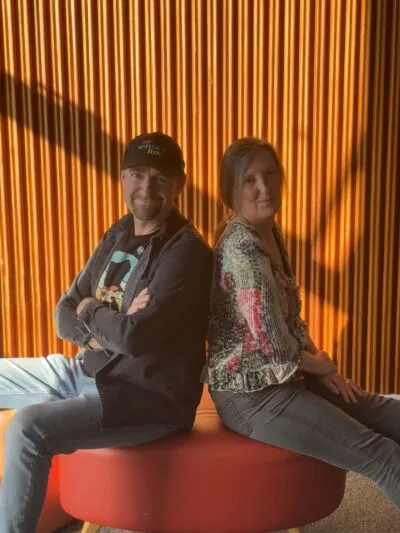 Stu and Debbie from No Ordinary Fish sitting back-to-back on a round bench against an orange wall, relaxed and friendly expressions, warm afternoon light.