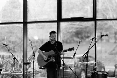 A black-and-white photo of Sam Jones playing an acoustic guitar and singing into a microphone indoors. The background is softly out of focus with windows letting in natural light.
