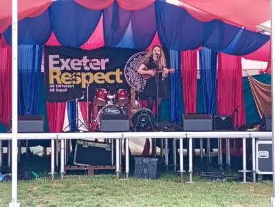 Cat Rose in black dress plays ukulele and sings on vibrant outdoor stage with “Exeter Respect” banner and drum kit.