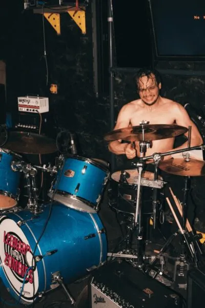 Drummer rocking out shirtless — Powerful image of a shirtless drummer mid-performance, sweat and energy filling the packed, intimate venue atmosphere.