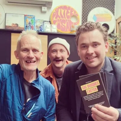 Rich Ellis, Ben, and Cameron indoors at Studio 54, Exeter, holding “Gig Buddies Exeter” flyers, surrounded by colourful artwork and music-themed decorations.